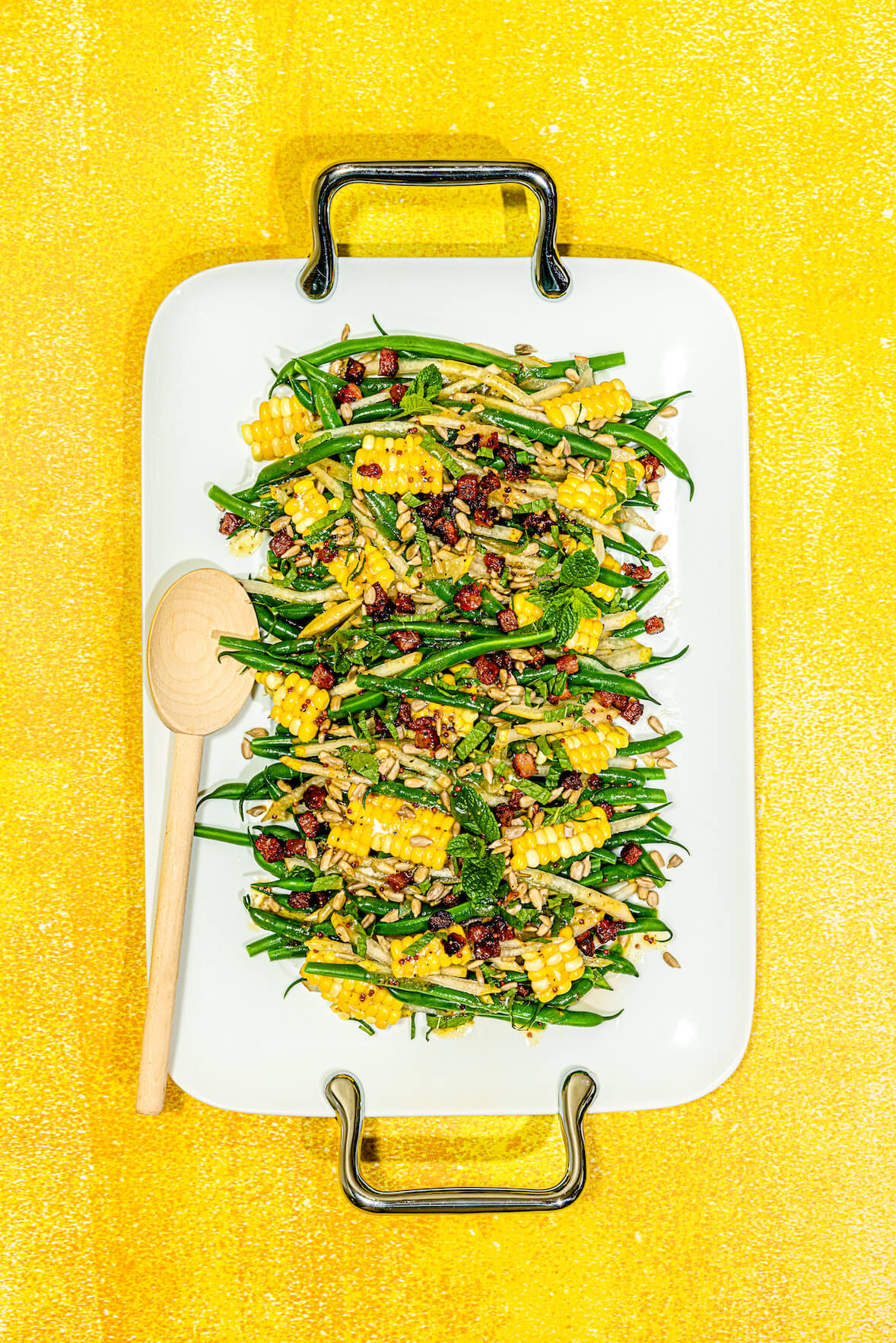 Salade de fèves vertes du jardin servies avec des poires coupées en juliennes et du maïs du marché avec de la pancetta. La salade est servie dans une assiette de service blanche avec deux poignées en métal.